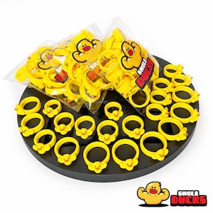 Peut inclure: Une collection d'anneaux en caoutchouc jaune vif en forme de canard disposés sur un présentoir circulaire noir. Certains anneaux sont libres, tandis que d'autres sont dans deux sachets en plastique transparent avec le texte Swole Ducks.