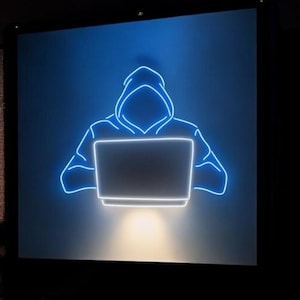 Könnte beinhalten: Ein Neonschild, das eine Person mit Kapuze zeigt, die einen Laptop benutzt. Die Umrisse der Figur und des Laptops leuchten hellblau vor einem dunklen Hintergrund. Der Laptop-Bildschirm strahlt ein weißes Licht aus.