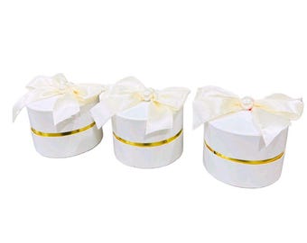 May include: Three white, round gift boxes with gold bands and ivory bows. Each bow is topped with a pearl. The boxes are arranged on a white background.