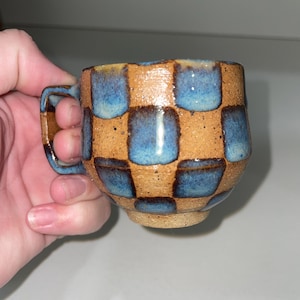 Op de afbeelding: Handgemaakte keramische mok met een bruin en blauw geblokt patroon. De mok heeft een klein handvat en een gespikkeld, getextureerd oppervlak. De binnenkant en het handvat zijn effen blauw. Een uniek stuk voor koffie of thee.