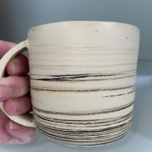 May include: Cream-colored ceramic mug with horizontal brown and black stripes. The mug has a rounded shape with a handle on the side. The stripes vary in thickness and spacing, creating a textured appearance. The mug is held by a hand.