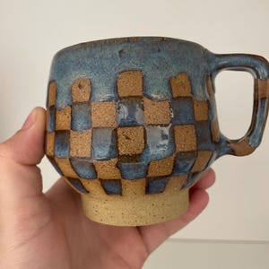 Op de afbeelding: Een keramische mok met een blauw en bruin geblokt patroon. De mok heeft een afgeronde vorm met een oor en een beige basis. De bovenkant van de mok is donkerblauw.