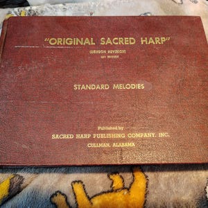 May include: A maroon-colored book titled "ORIGINAL SACRED HARP" with gold lettering. The cover also reads "STANDARD MELODIES" and "SACRED HARP PUBLISHING COMPANY, INC. CULLMAN, ALABAMA."