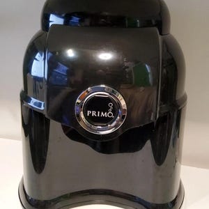 Może przedstawiać: Czarny dystrybutor wody Primo z błyszczącym wykończeniem. Dystrybutor ma zaokrąglony kształt ze srebrnym okrągłym emblematem z napisem "PRIMO". Dystrybutor jest przeznaczony do przechowywania i dozowania wody.