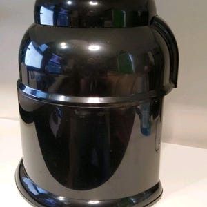 Może przedstawiać: Czarny plastikowy pojemnik z błyszczącym wykończeniem. Pojemnik ma cylindryczną podstawę, zaokrągloną środkową część i mniejszą, zaokrągloną g&oacute;rę. Zakrzywiony uchwyt jest przymocowany do boku. Pojemnik wydaje się być dzbanem do chłodziarki wody.