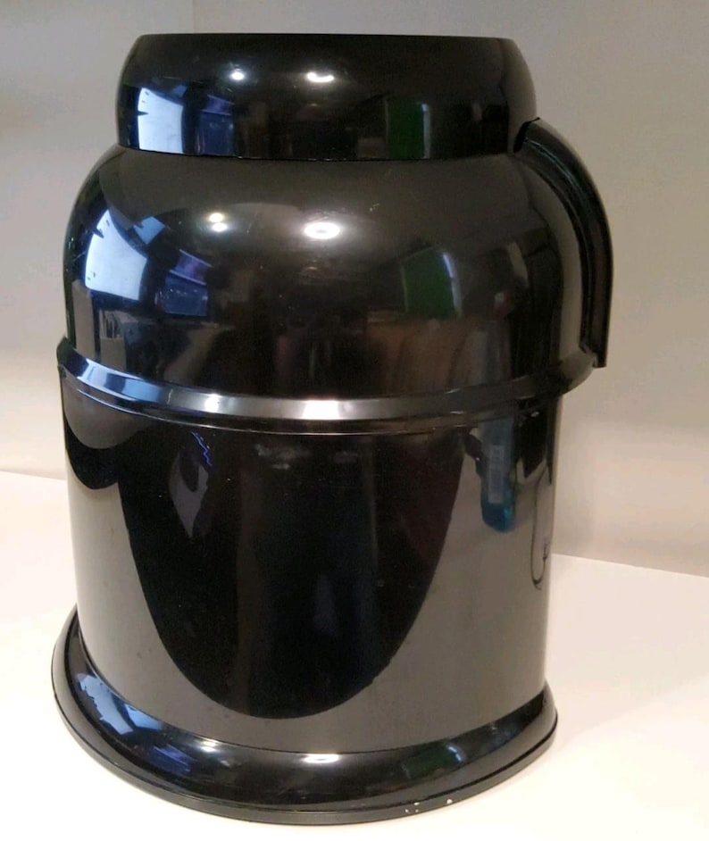 Może przedstawiać: Czarny plastikowy pojemnik z błyszczącym wykończeniem. Pojemnik ma cylindryczną podstawę, zaokrągloną środkową część i mniejszą, zaokrągloną g&oacute;rę. Zakrzywiony uchwyt jest przymocowany do boku. Pojemnik wydaje się być dzbanem do chłodziarki wody.