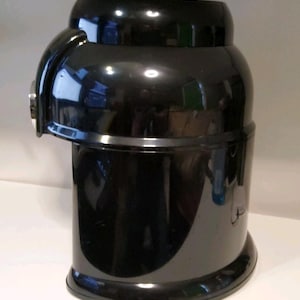 Może przedstawiać: Błyszczące czarne urządzenie kuchenne o zaokrąglonym korpusie i zakrzywionym uchwycie z boku. Urządzenie ma cylindryczny kształt z lekko rozszerzoną podstawą i płaskim wierzchem. Powierzchnia odbija światło.