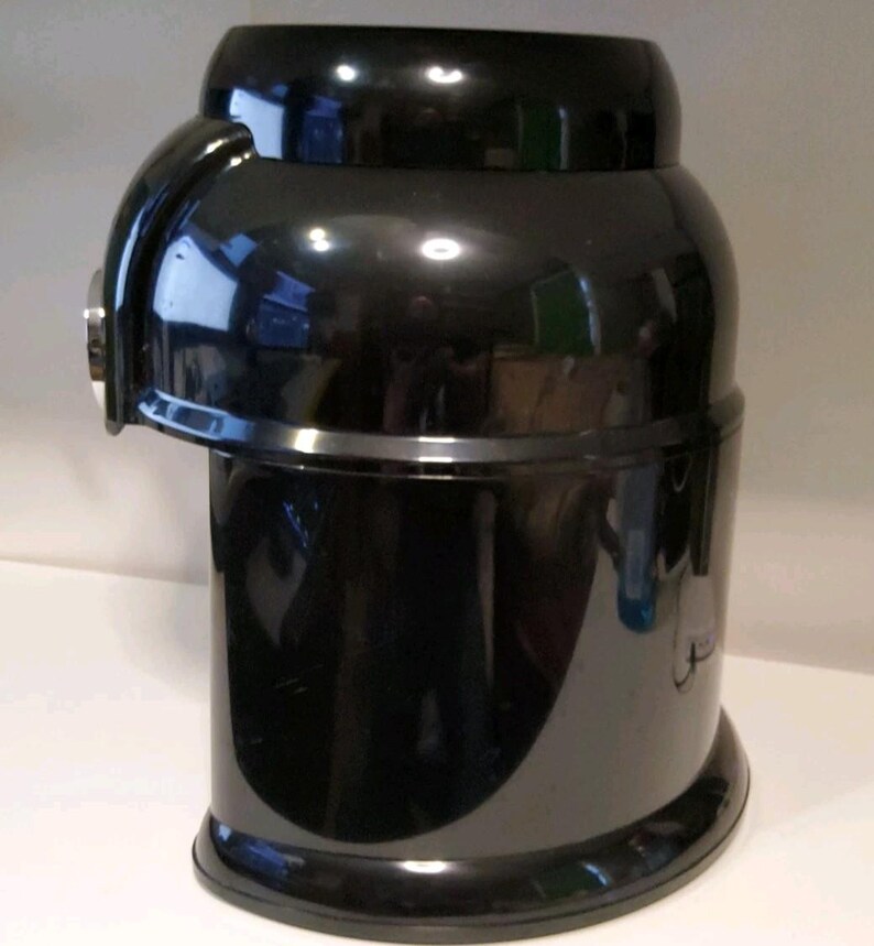 Może przedstawiać: Błyszczące czarne urządzenie kuchenne o zaokrąglonym korpusie i zakrzywionym uchwycie z boku. Urządzenie ma cylindryczny kształt z lekko rozszerzoną podstawą i płaskim wierzchem. Powierzchnia odbija światło.