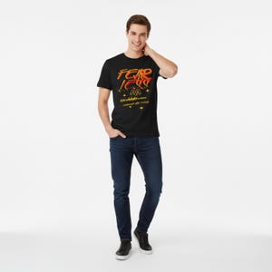 Puede incluir: Camiseta negra con el texto "FEAR LESS" en naranja y amarillo, con texto adicional y gráficos de estrellas. La camiseta la lleva una persona con vaqueros azul oscuro y zapatos negros.