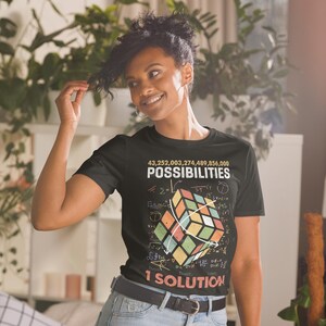 Peut inclure: T-shirt noir avec un graphique de cube de Rubik coloré et le texte "43,252,003,274,489,856,000 POSSIBILITIES 1 SOLUTION". Le t-shirt présente des équations mathématiques autour du cube.