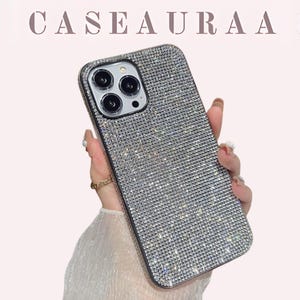 以下が含まれることがあります： キラキラ光るラインストーンで覆われたシルバーの携帯電話ケース。ケースには黒い縁とカメラの切り抜きがあります。「CASEAURA」の文字が画像上部にあります。ケースは人が持っています。