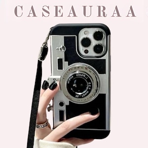 May include: A black and silver phone case designed to resemble a vintage camera. The case has a lens detail, a black strap, and the text "CASEAURA" at the top. The phone case is held by a person with black nail polish.