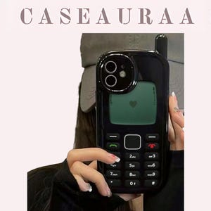 May include: Black phone case with a retro cell phone design. The case features a screen with a heart icon and a keypad with numbers and symbols. The brand name "CASEAURA" is displayed at the top. The case is held by a person.