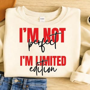 Puede incluir: Sudadera color crema con el texto "I'M NOT perfect I'M LIMITED edition" en rojo y negro. La sudadera está doblada, con unos vaqueros azules visibles en la parte inferior. Una pequeña planta suculenta en una maceta blanca está en el fondo.