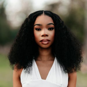 May include: A portrait of a person with dark skin, wearing a white halter top. The person has long, curly black hair and is looking directly at the camera. They have a nose ring and dark lipstick. The background is blurred with green and brown tones.