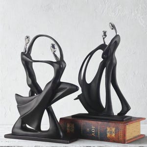 Könnte beinhalten: Zwei abstrakte schwarze Skulpturen, die Figuren in anmutigen Posen darstellen. Die Skulpturen haben silberne Akzente auf den Köpfen und sind vor einem weißen Hintergrund platziert. Eine Skulptur steht auf einem rechteckigen Sockel, die andere auf einem alten Buch.