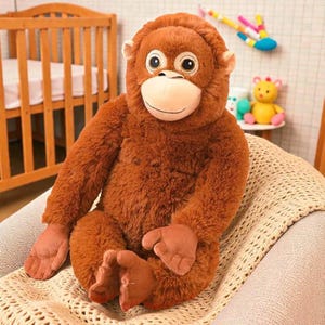 May include: A plush orangutan toy with brown fur, large eyes, and a tan face. The stuffed animal is sitting on a cream-colored blanket. A wooden crib is visible in the background.