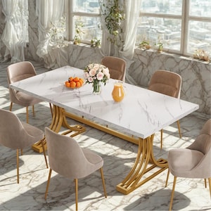 Puede incluir: Una mesa de comedor rectangular con una tapa blanca con aspecto de mármol y una base dorada curvada. Seis sillas tapizadas beige rodean la mesa. Un jarrón de flores, un cuenco de naranjas y una jarra de zumo están sobre la mesa.