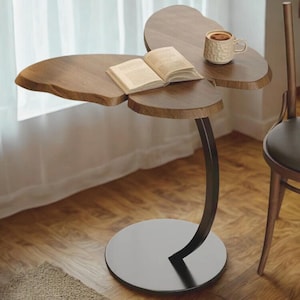 Może przedstawiać: Unikalny, trzydzielny drewniany stolik boczny z ciemną metalową podstawą. Stół ma naturalne wykończenie słojów drewna i zawiera otwartą książkę i kubek. Stół ma około 76 cm wysokości.