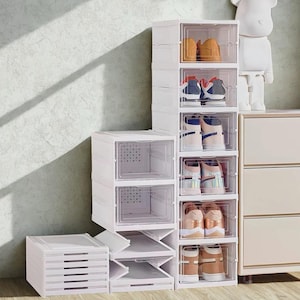 May include: White plastic shoe storage containers with clear front doors. Several pairs of shoes are visible inside the containers. A stack of drawers is on the left. A white bear figurine is on a shelf in the background.