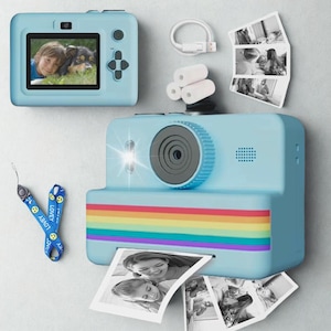 Puede incluir: Cámara instantánea azul claro con una franja de arcoíris y una foto impresa. La cámara tiene una lente, flash y una pequeña pantalla que muestra una foto de un niño y un perro. Los accesorios incluyen una correa para la muñeca y un cable de carga.