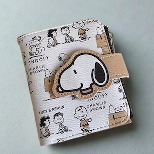 Puede incluir: Una cartera blanca con Snoopy y otros personajes de Peanuts. Tiene una aplicación beige de Snoopy en la parte delantera y un cierre de correa beige. Las palabras "Snoopy" y "Charlie Brown" están impresas.