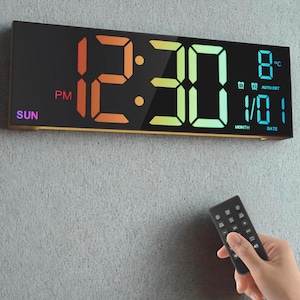 Może przedstawiać: Prostokątny zegar cyfrowy wyświetla godzinę 12:30 PM, z dniem tygodnia, datą i temperaturą. Zegar ma czarną tarczę z wielokolorowymi cyframi LED i pilotem.