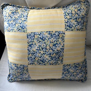 May include: A square throw pillow featuring a patchwork design of alternating fabric blocks. The pattern consists of light yellow fabric with thin white and blue stripes, contrasted with a blue and yellow floral print fabric in a grid layout.