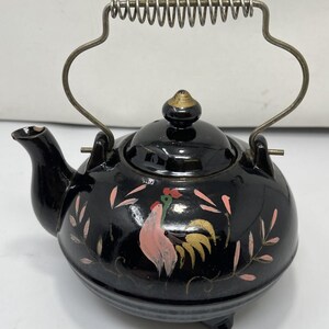 Pode incluir: Um bule de cerâmica preta com um design de galo pintado à mão em tons de rosa, amarelo e verde. O bule tem uma tampa a condizer com um pequeno puxador e uma pega de arame metálico com mola. O bico apresenta uma pequena lasca na ponta.