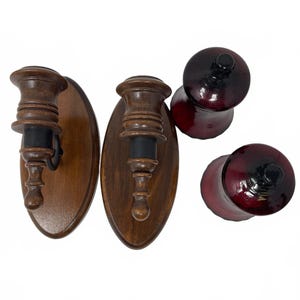 May include: Two wall-mounted candle holders with oval wooden backplates and turned wooden sconces featuring black accents, paired with two separate deep red glass candle cups with black rims, shown from a top-down perspective.