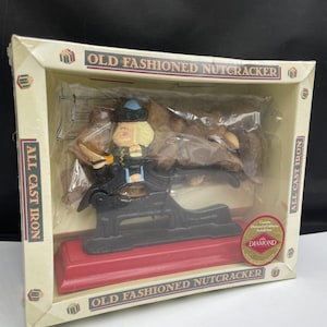 May include: An old fashioned nutcracker made of black cast iron with a red base, packaged in a box labelled "OLD FASHIONED NUTCRACKER" and "ALL CAST IRON". The nutcracker features a painted figure with a blue hat and yellow hair.