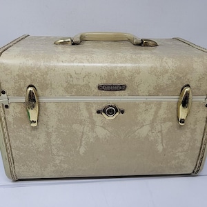May include: A vintage off-white Samsonite train case with a textured, mottled finish. The rectangular hard-shell case features a top handle, two gold-tone metal latches, and a central locking mechanism. The brand name Samsonite is embossed on a small metal plate.