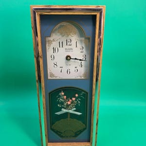 May include: A rectangular mantel clock with a light wood frame and chrome trim. The clock face features black Arabic numerals, the text Elgin Clock Co, and a floral design. Below the face is a dark green panel with a floral bouquet and ribbon.