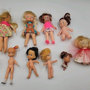 May include: A collection of nine small plastic dolls with various hair colors and styles. Some dolls are wearing miniature dresses, skirts, or tops with floral and geometric patterns, while others are unclothed. One doll wears a red tulle skirt.