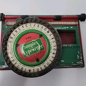 以下が含まれることがあります： 文字と数字が記された円形のダイヤルを備えたヴィンテージのSimplex Typewriter Number 100。赤と緑の金属製フレームには塗装の剥がれが見られます。中央の赤と緑のラベルには「The Simplex Typewriter」というテキストが表示されています。