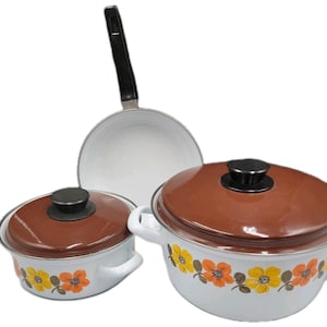 Puede incluir: Un juego de utensilios de cocina de esmalte blanco que incluye dos ollas con tapas marrones y una sartén con mango negro. El exterior de las ollas está decorado con un patrón repetitivo de flores naranjas y amarillas con hojas verdes.