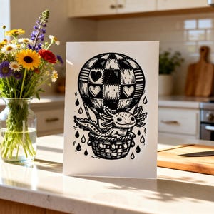 May include: Black and white illustration of an axolotl in a hot air balloon with heart-shaped patches. The axolotl is in a woven basket, with raindrops falling around it. The balloon has a checkered pattern.