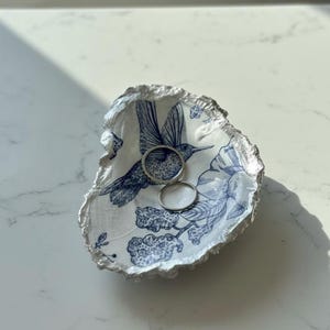 May include: A decorative oyster shell dish with a blue hummingbird and floral design. The rim is silver, and two silver rings rest inside the dish. The dish is on a white surface.