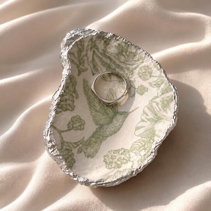 May include: A small, shell-shaped trinket dish with a cream-colored interior featuring a green botanical print of a hummingbird and flowers. The edges of the dish are finished with a textured silver metallic trim. A simple silver ring rests inside.