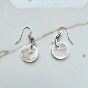 May include: A pair of earrings featuring a circular, iridescent, mother-of-pearl design. The earrings have silver-colored fish hook ear wires. The earrings are displayed on a white and gray marble surface.