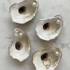 May include: Four oyster shells used as small dishes, each featuring a smooth white interior with a dark brown center spot. Three shells have silver-leafed edges and one white pearl, while the fourth has a gold-leafed edge, a pearl, and a gold ring.