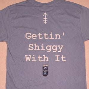 May include: A light blue t-shirt with the phrase "Gettin' Shiggy With It" in white block letters. A small can of beer is printed below the text. A white arrow and symbol are printed above the text.