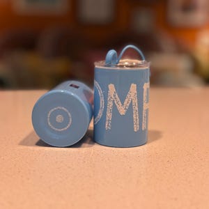 May include: Two light blue cylindrical containers with white text that reads "OMH". One container is upright, the other is on its side. The upright container has a small handle and a lid. The bottom of the containers have a silver design.