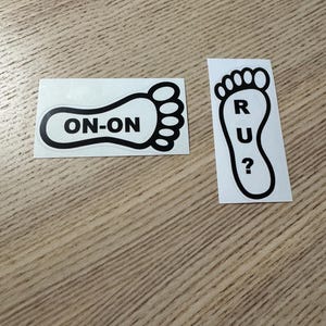May include: Two black and white stickers in the shape of feet. One sticker has the text "ON-ON" inside the foot outline. The other sticker has the letters "R U?" inside the foot outline.