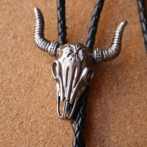 May include: A bolo tie featuring a silver-toned metal bull skull pendant with detailed horn textures and a floral motif on the forehead. The pendant is attached to a black braided leather cord that runs through the back of the skull structure.
