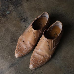 May include: A pair of light brown leather slip-on shoes with decorative stitching on the toe and sides. The shoes have a pointed toe and elastic side panels. The shoes are approximately 10 inches long.