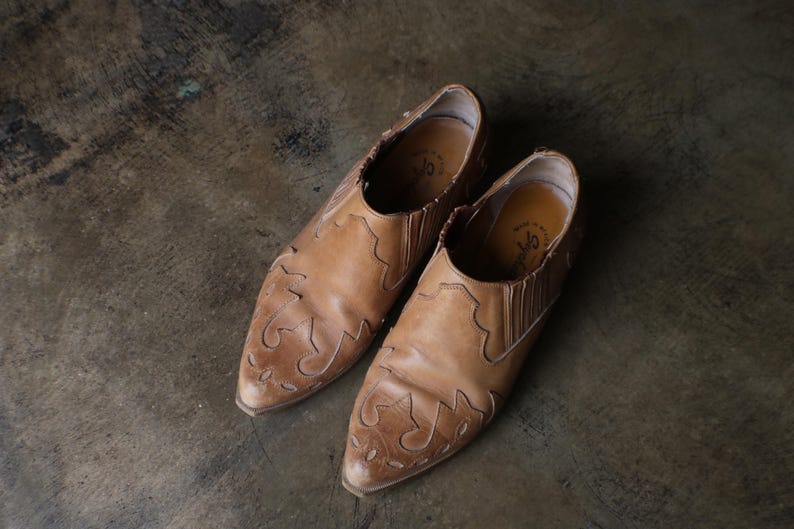 May include: A pair of light brown leather slip-on shoes with decorative stitching on the toe and sides. The shoes have a pointed toe and elastic side panels. The shoes are approximately 10 inches long.