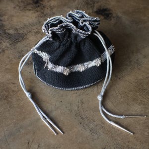 May include: A small, round, black drawstring pouch crafted from knit fabric. The bag features a silver-toned metallic trim around the top opening and a horizontal band decorated with small, silver-colored leaf-shaped embellishments.