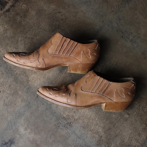 May include: A pair of tan leather ankle boots with pointed toes and decorative cutouts. The boots have a low heel and a Western-inspired design. The boots are displayed on a neutral, textured surface.
