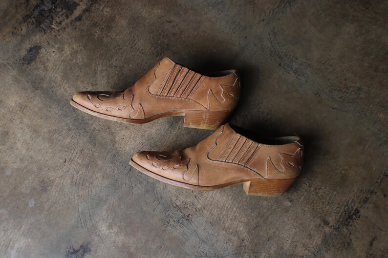 May include: A pair of tan leather ankle boots with pointed toes and decorative cutouts. The boots have a low heel and a Western-inspired design. The boots are displayed on a neutral, textured surface.